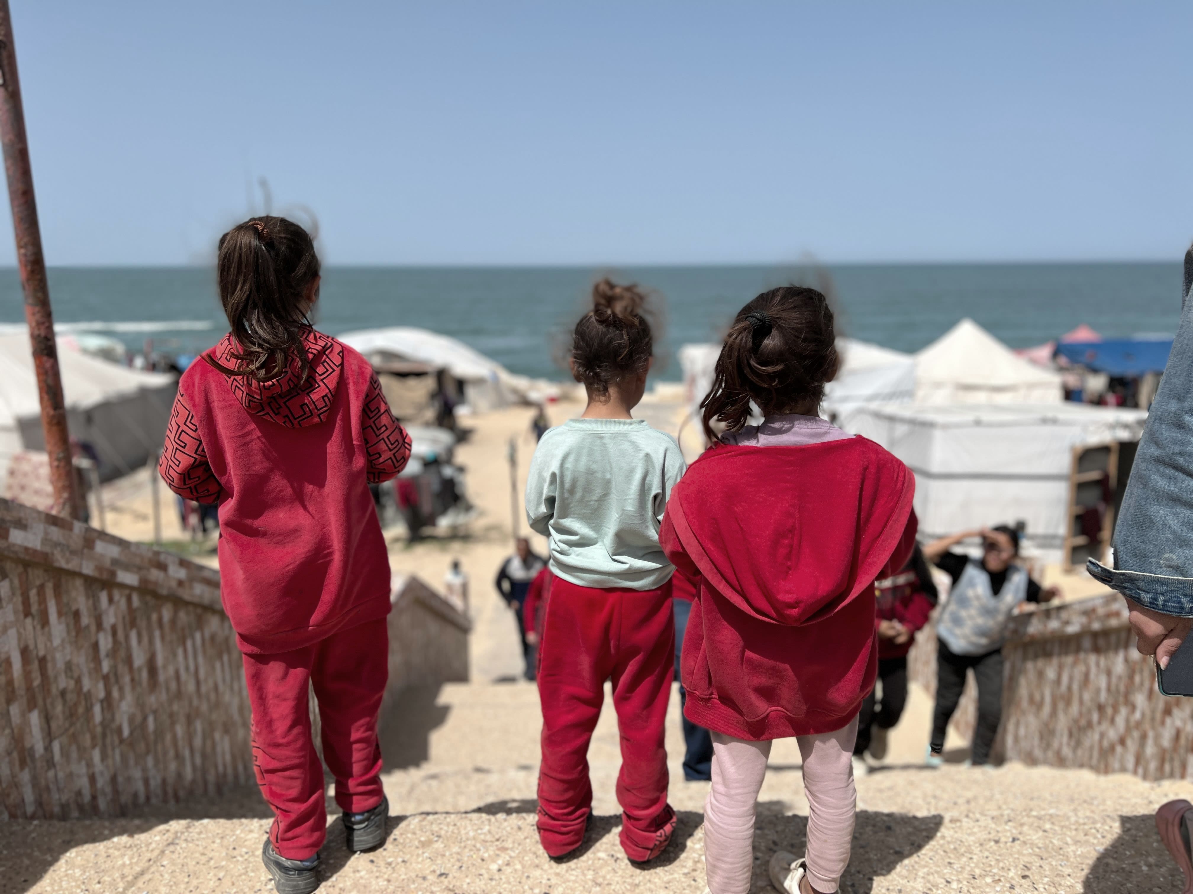 Three young girls dressed in red look out towards the sea in Rafah, Gaza.