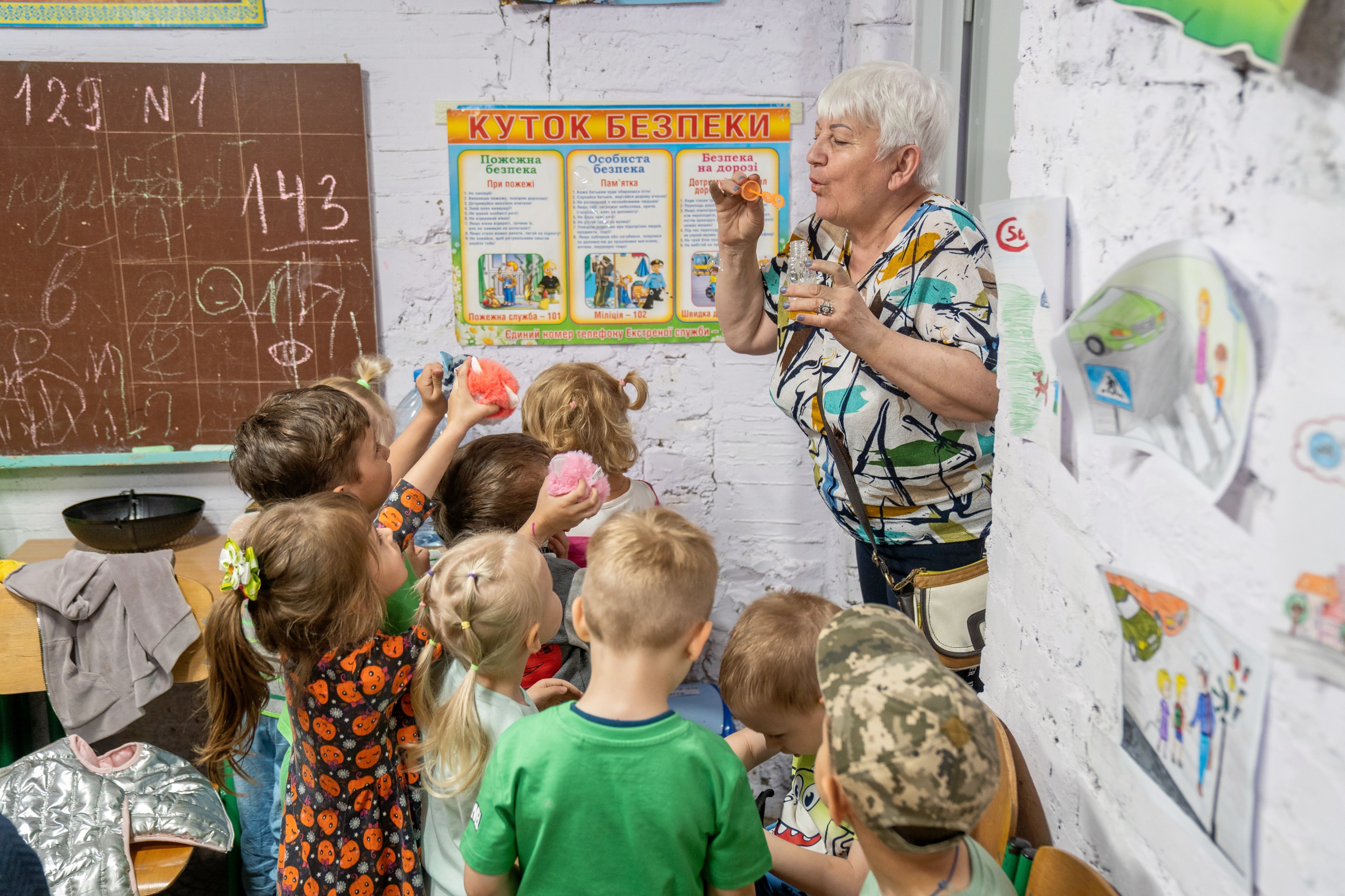 A teacher blows soap bubbles playing with children in the kindergarten shelter during an air raid.