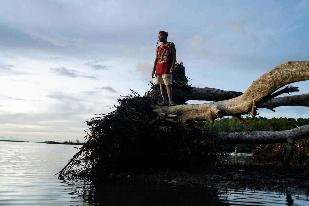 Photo: George, 15, wears a red vest and looks out at the water while standing on a tree that was knocked down during a storm in a community affected by rising sea levels in the Solomon Islands. Credit: Conor Ashleigh / Save the Children.