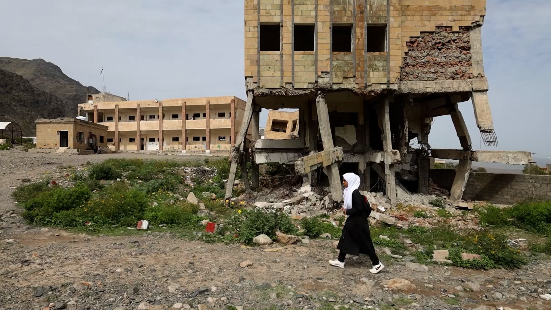 Lina*, 12, walks around the grounds of her semi-destroyed school that's contaminated with landmines in Taiz, Yemen. She wears a white headscarf and the building immediately behind her is shown partly destroyed. A safer, more complete school building is shown in the background.