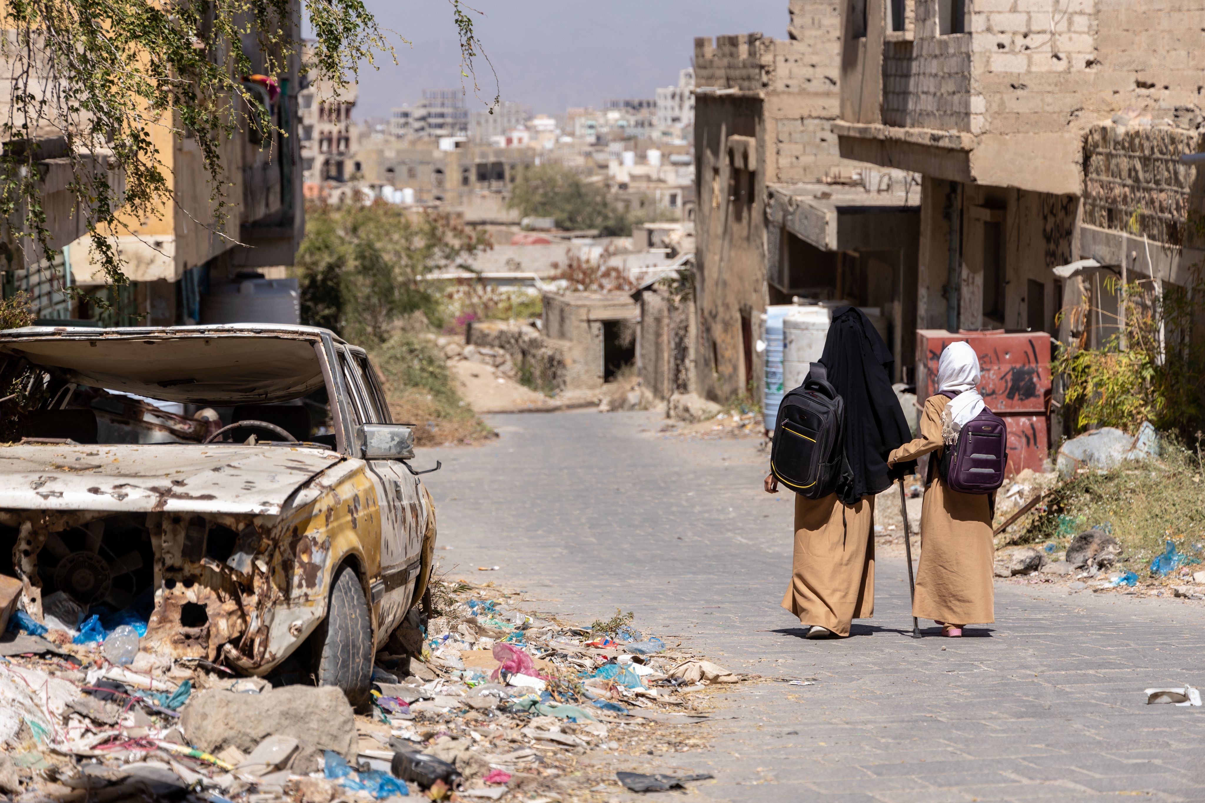 Maha, 10, & Maya, 16, walk home from school, passing destroyed vehicles and buildings in Yemen