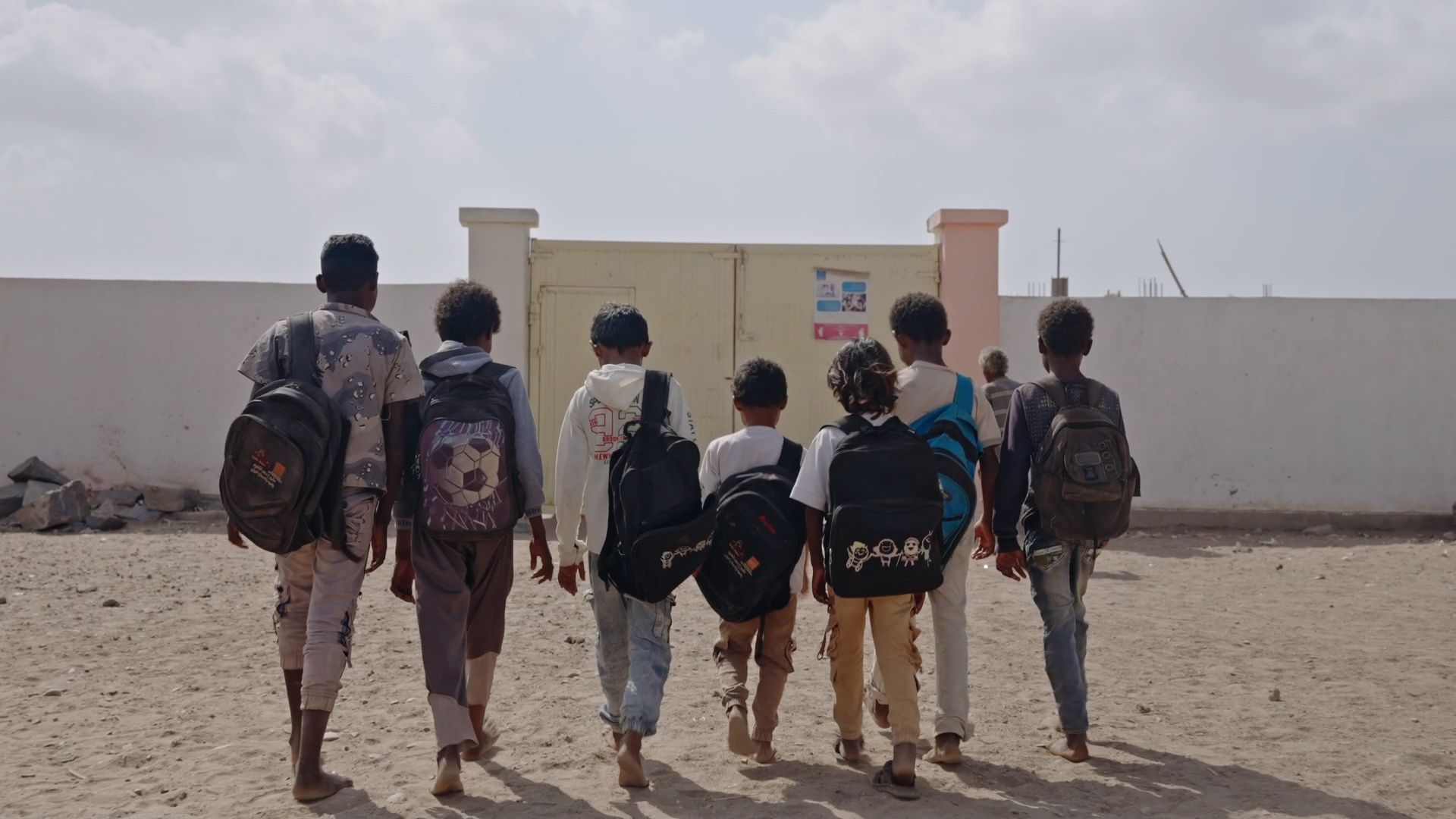 A group of children carrying backpacks make their way toward a large gate at a school in Lahj, Yemen